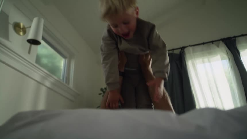 A father lifts his blonde son over his head with his hands and falls back onto a bed