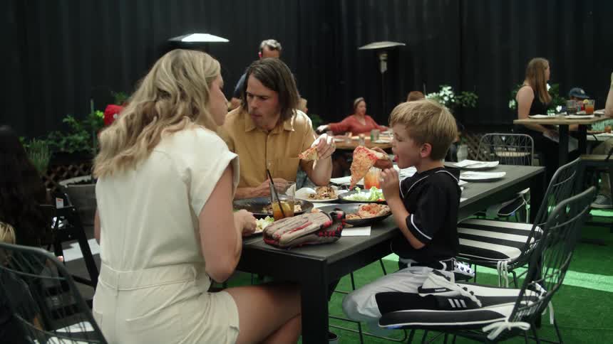A blonde child sits at a table eating pizza next to his blonde mother and father