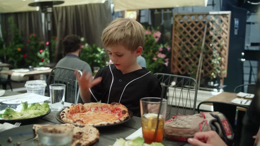 A blonde boy sits next to his blonde mother while he pokes his pizza smiling