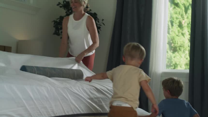 A boy helps his mother pulls sheets on the bed while the younger boy pulls a mat off the sheets