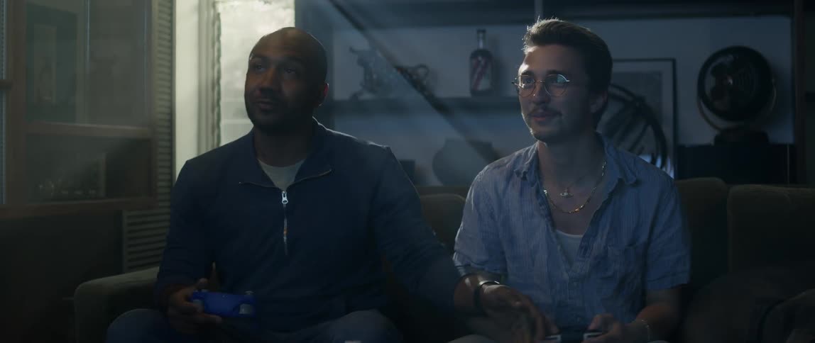 Two men are sitting on a couch holding remote controllers and laughing as they play video games