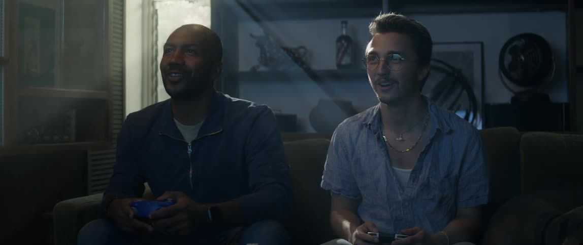A pair of men play video games together on a sofa in a dimly lit living room