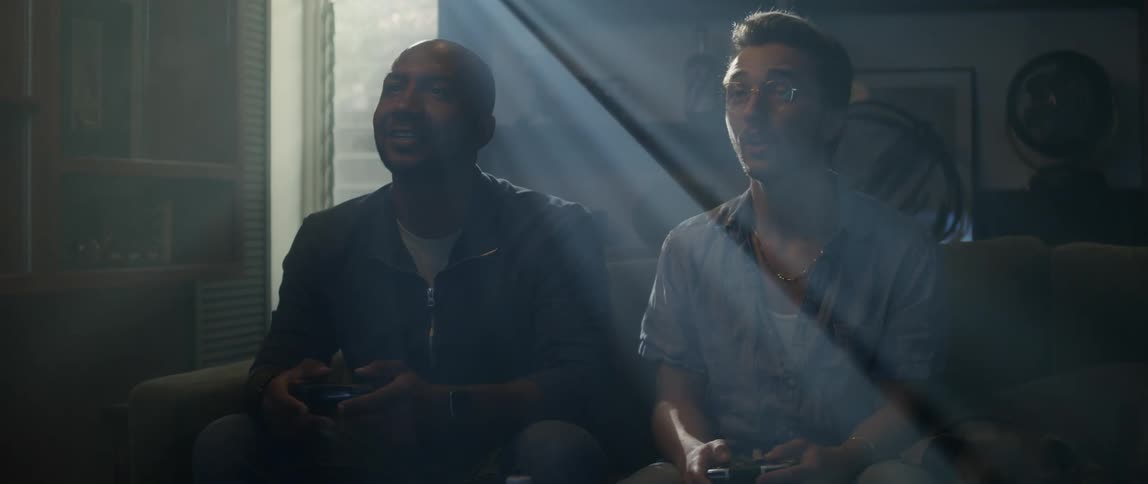 Two men sit on a couch and laugh as they play a video game together