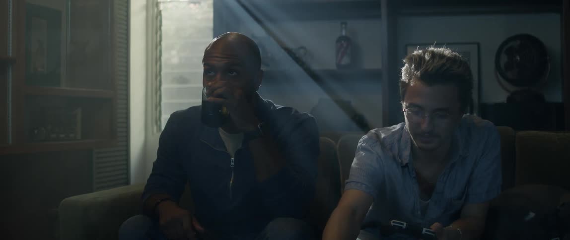 Two men are sitting on a couch holding remote controllers for a video game and having chips and pop