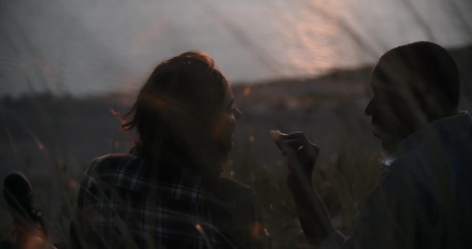 A man feeds a woman a bite of food while enjoying an ocean side picnic at dusk