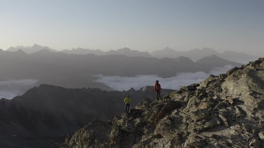 A wide open view of a mountain range is shown from the top via drone with two people standing on one of the mountains