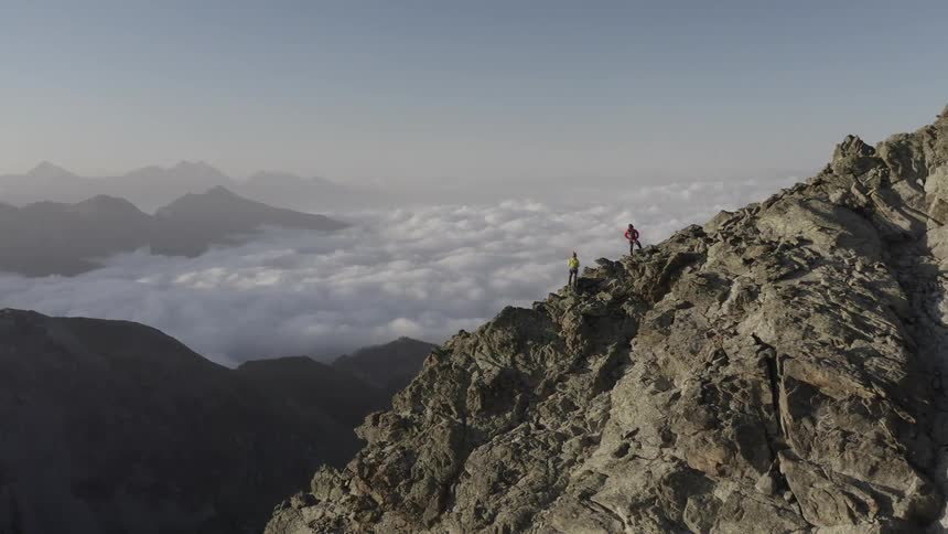 Two people are standing high on a mountain overlooking clouds and more mountains in the distance