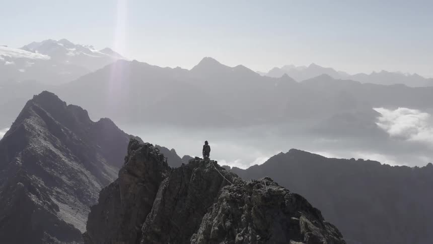 A mountaineer walks across the top of a narrow mountain ridge while unrolling a rope
