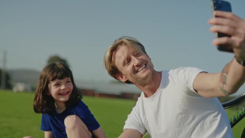 A man uses his smartphone to take a selfie with a child sitting with him in a field during the day