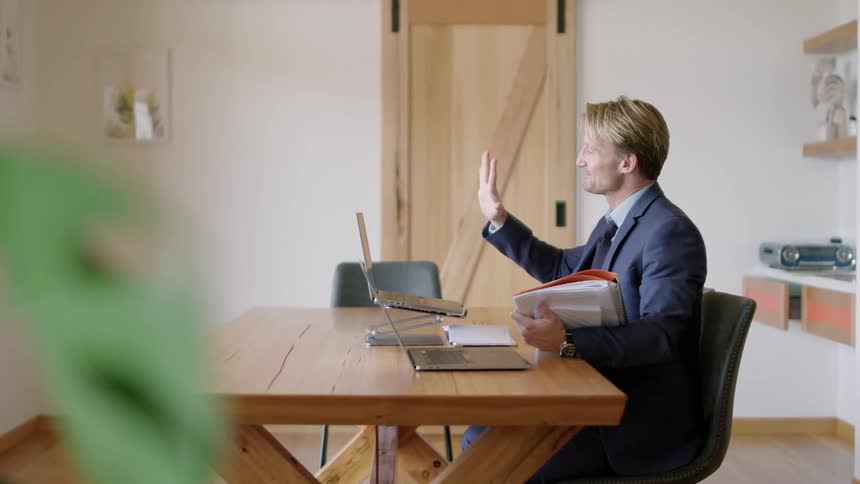 Man sitting at table waves goodbye and ends video call on laptop before turning to paperwork