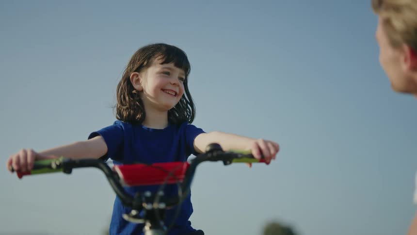 A little girl is sitting on her bicycle holding the handlebars and smiling as she talks to her father
