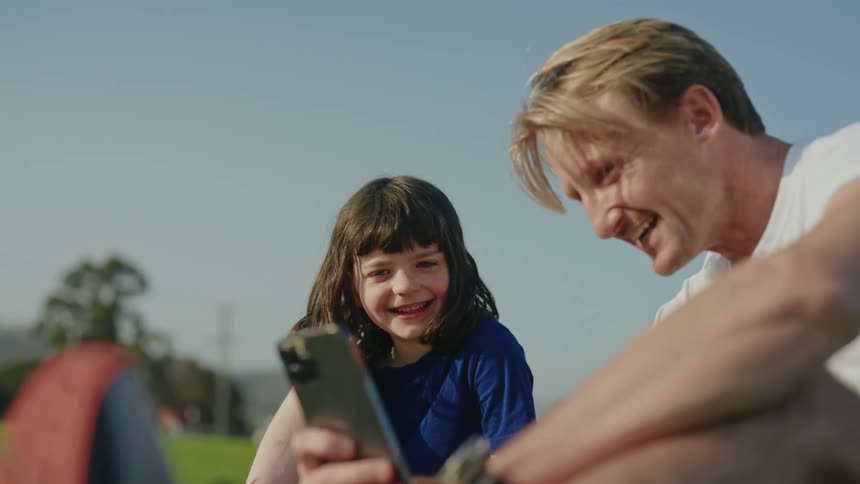 A man sits on the grass and laughs as he shows his daughter his cell phone