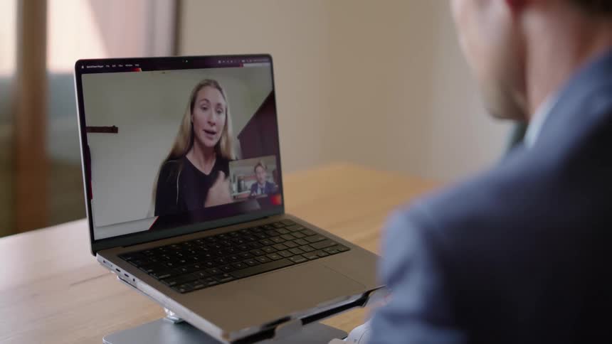 A man in a suit is sitting at a desk having a video conference with a woman on a laptop computer