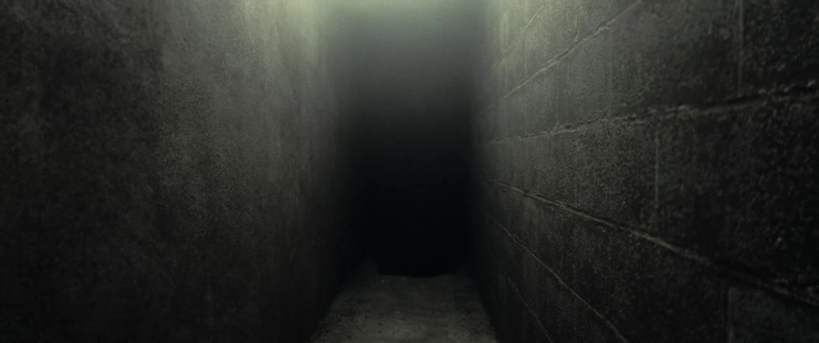 A person wearing a long black robe and hood is walking down a long and narrow corridor with cinder block walls