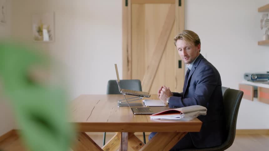 A man in a suit is sitting at a desk having a video conference on a laptop computer and he picks up a notebook from the desk