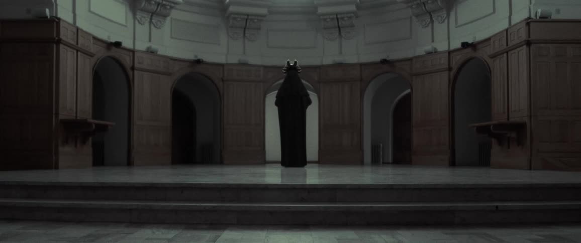 A cloaked figure is standing on a stage with many arches leading out