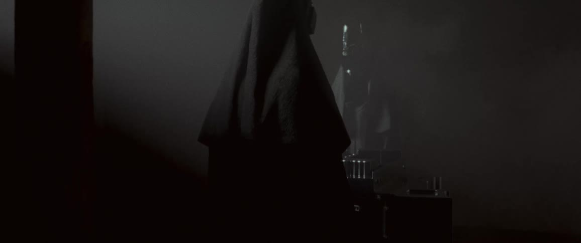 Two people wearing tribal masks and hoods face each other in foggy dim room
