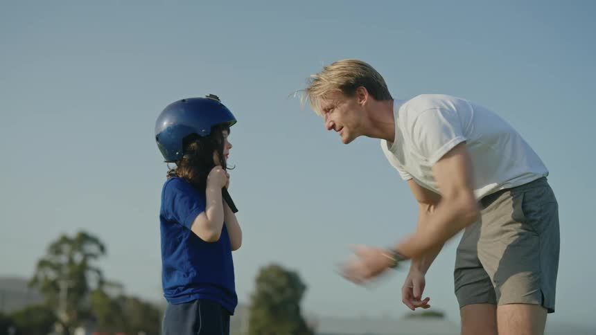 A man is standing outside with his young daughter as he puts a cycling helmet on her head and she fastens the strap under her chin and they walk to her bicycle