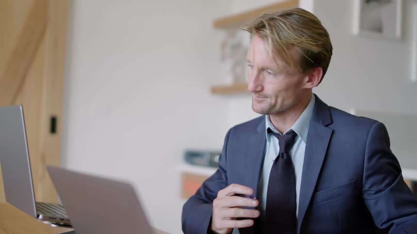 A man sits in his office and talks with someone over his laptop as she looks at a binder and writes down information