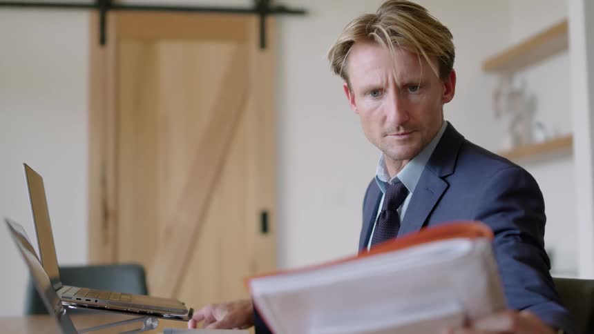 Businessman sits at desk working on paperwork and using laptop