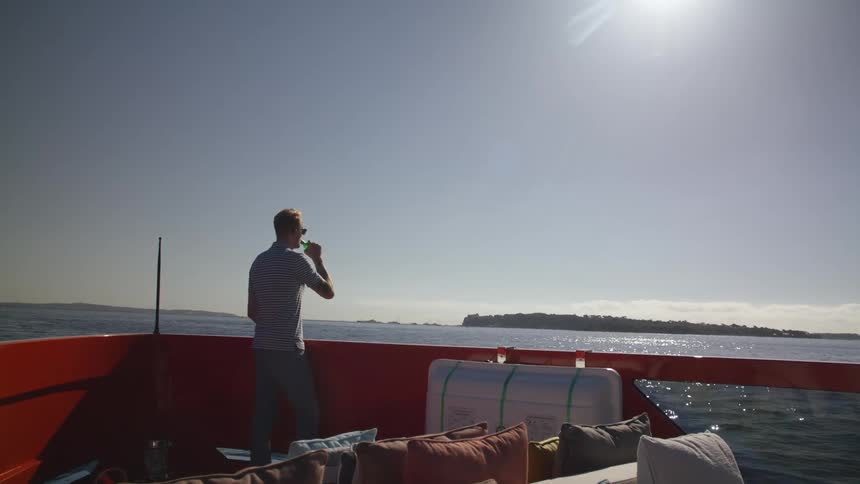 Man wearing sunglasses and in gray shirt and jeans drinks from green bottle on boat traveling on ocean on a sunny day