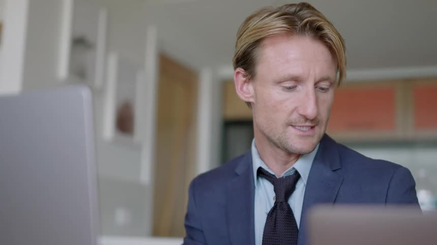 A man in a suit is sitting in front of a laptop computer smiling as he has a video conference and waves goodbye