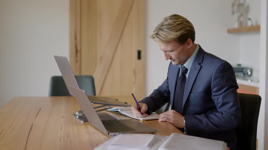 A man in a suit is sitting at a desk with a laptop computer and holding a pen as he looks though a book