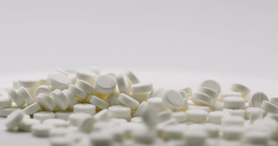 A pile of white pills are spread across a table and more pills fall into the pile from above