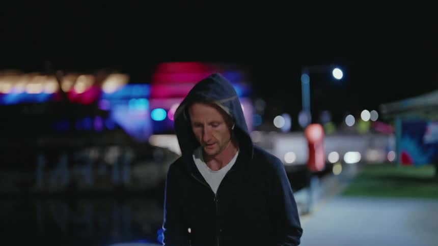 A man next to a river puts ear buds in and lifts his hood up and starts to jog