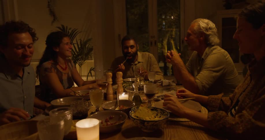 A group of five adults sit around the table eating dinner and conversing clearly laughing and having a good time with one another