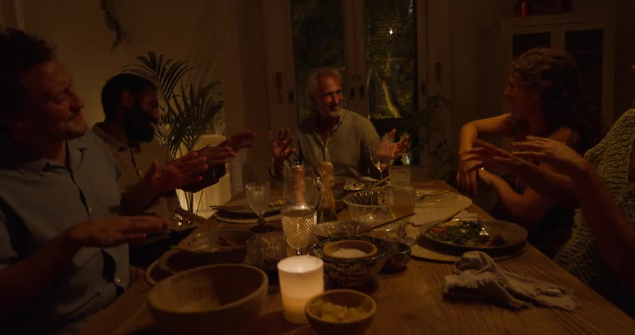 A group of men and women sit at a table in a home and move their arms around as they dance and laugh