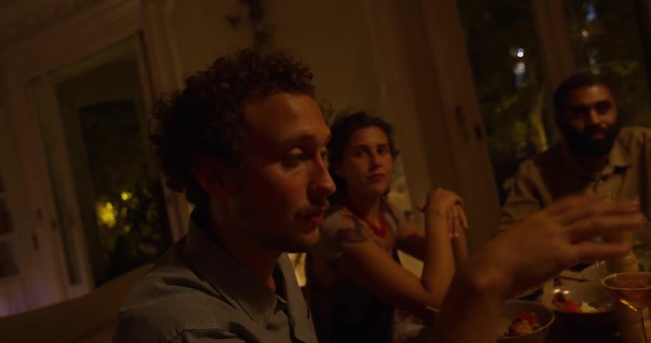 Man and woman listen and watch as gesturing man talks during dinner party