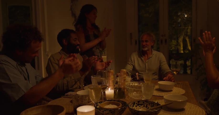 Friends sitting around table at night clap as older man blows out birthday candle