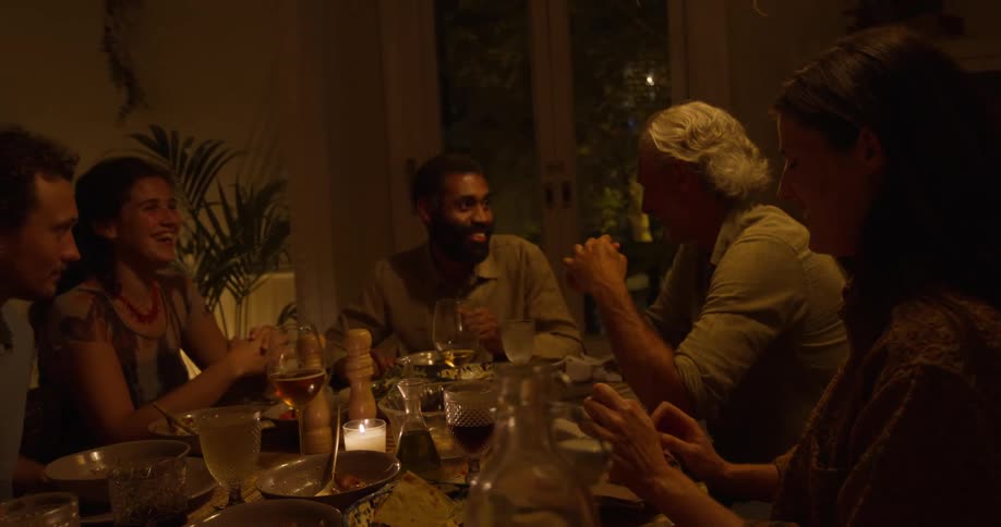 A group of people chat happily while eating together in a home dining room at night