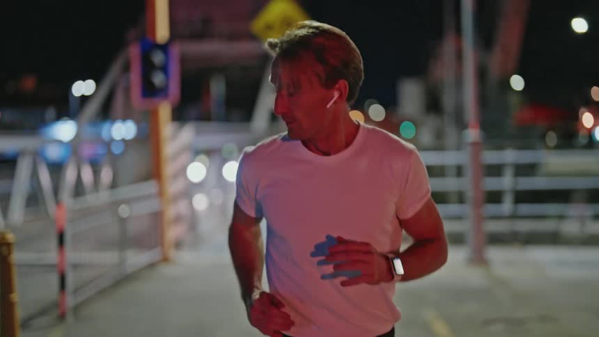 Blonde man with earphones white t shirt and black shorts runs over bridge at night