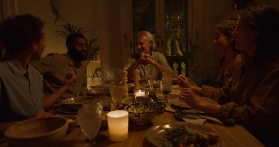 A group of friends sit at a dining table in a home and talk and laugh together during a meal