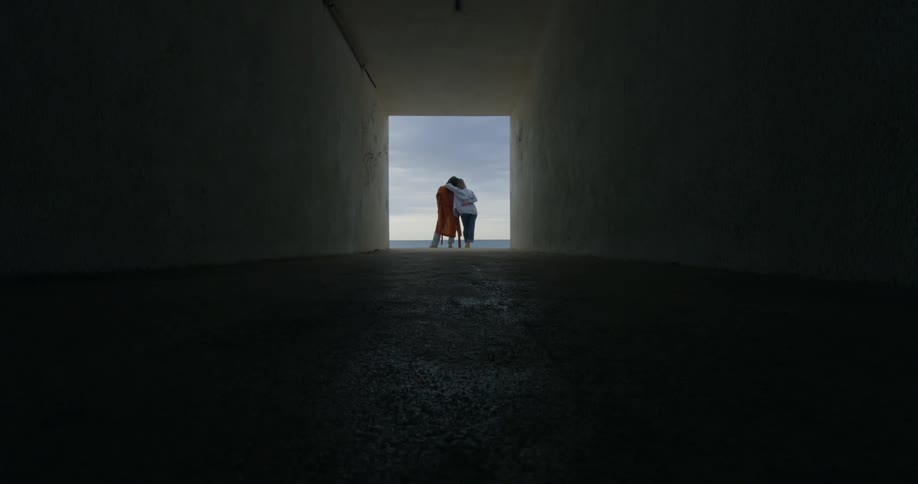 Two women standing at the square opening of a tunnel hugging as they look at the ocean