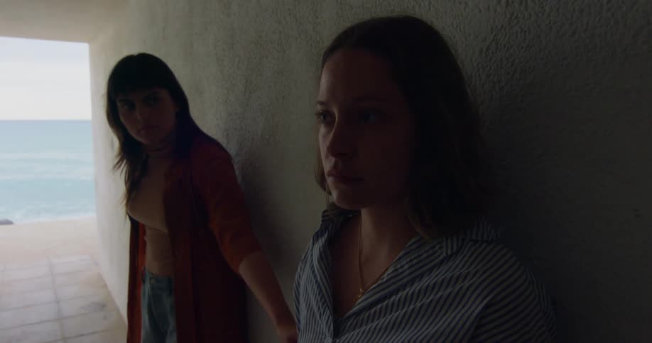 Woman with red coat orange top and jeans touches womans arm with brown hair standing in corridor overlooking ocean