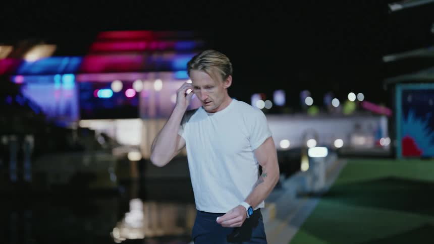 Blonde man in white shirt warms up and wears earphones outdoors at night