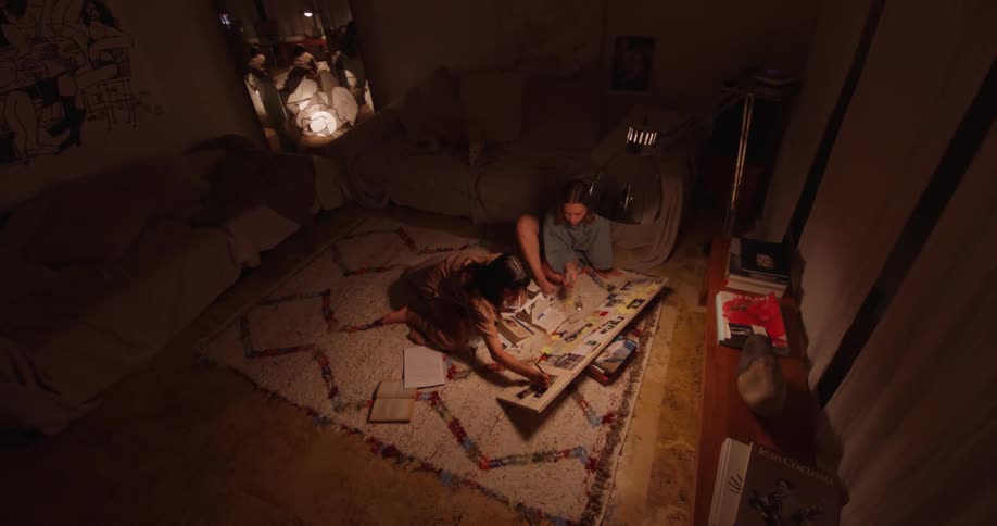 Two women sit on carpet working on research together in a dim room