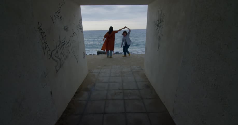 Two women dance on ground overlooking beach at end of graffiti decorated concrete tunnel