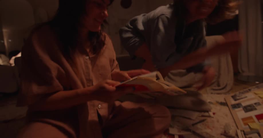 Two women wearing pajamas sits on floor looking at photos and books in room