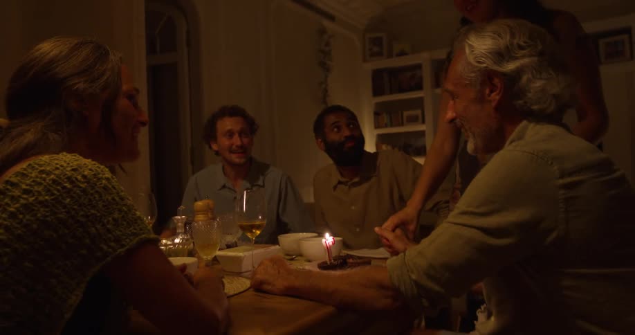 Man with gray hair and shirt blows out birthday candles sitting at dining table with friends and wife