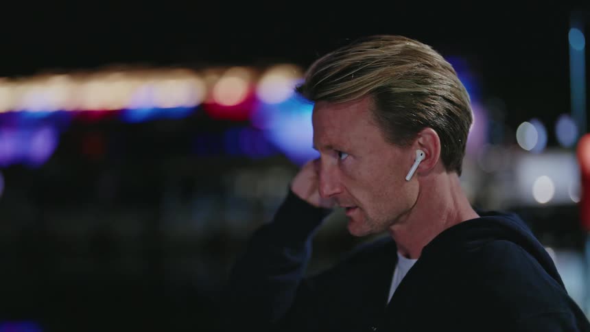 A man looks at his watch puts in his earbuds and puts the hood of his jacket on and starts to run