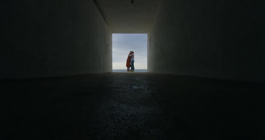 Two women stand at the end of a tunnel on a beach and hug one another