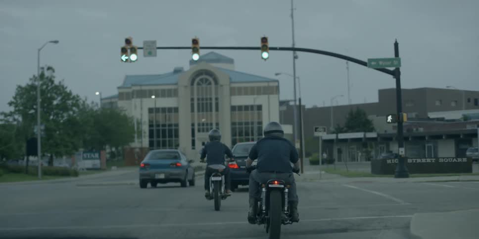 Two people riding motorcycles follow behind two cars on city street at dawn