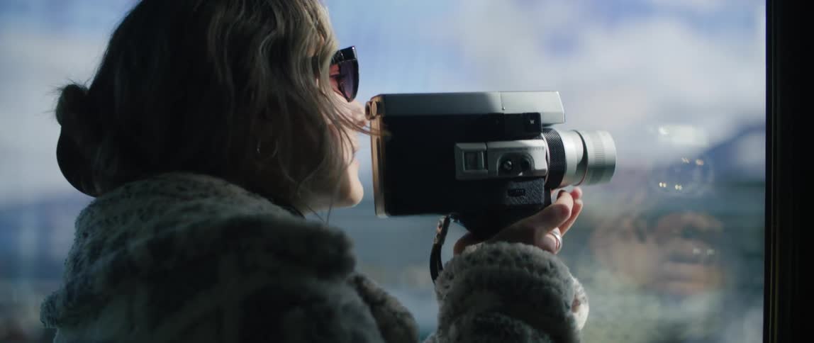 Woman wearing coat stands near reflective window videotaping through the window