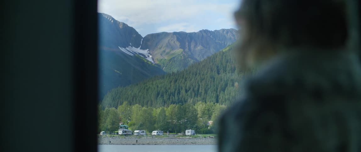 An elderly woman gazes out a window at a mountain range and pine trees