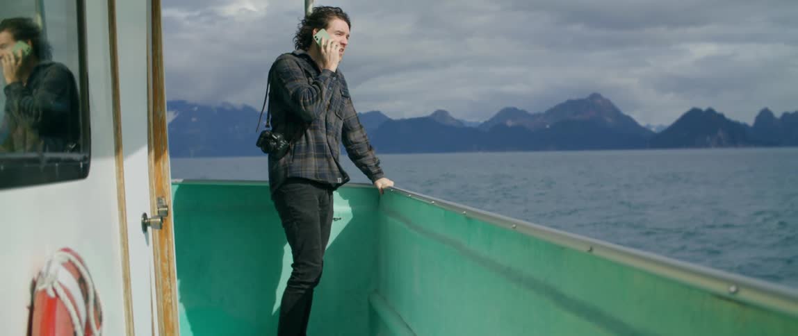 Man walks out onto boat deck to stand near rail while talking on the phone with foggy mountainous shore in the background