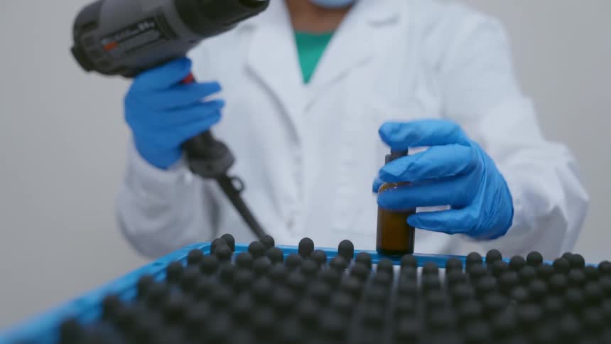 A lab worker uses a dryer to shrink wrap dropper bottles